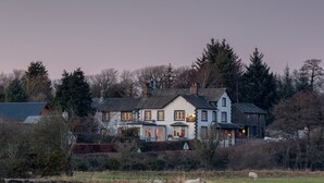 A large, two-story building with a traditional architectural style, surrounded by a rural landscape with fields and trees. - Lutwidge Arms Limited (Holmrook)