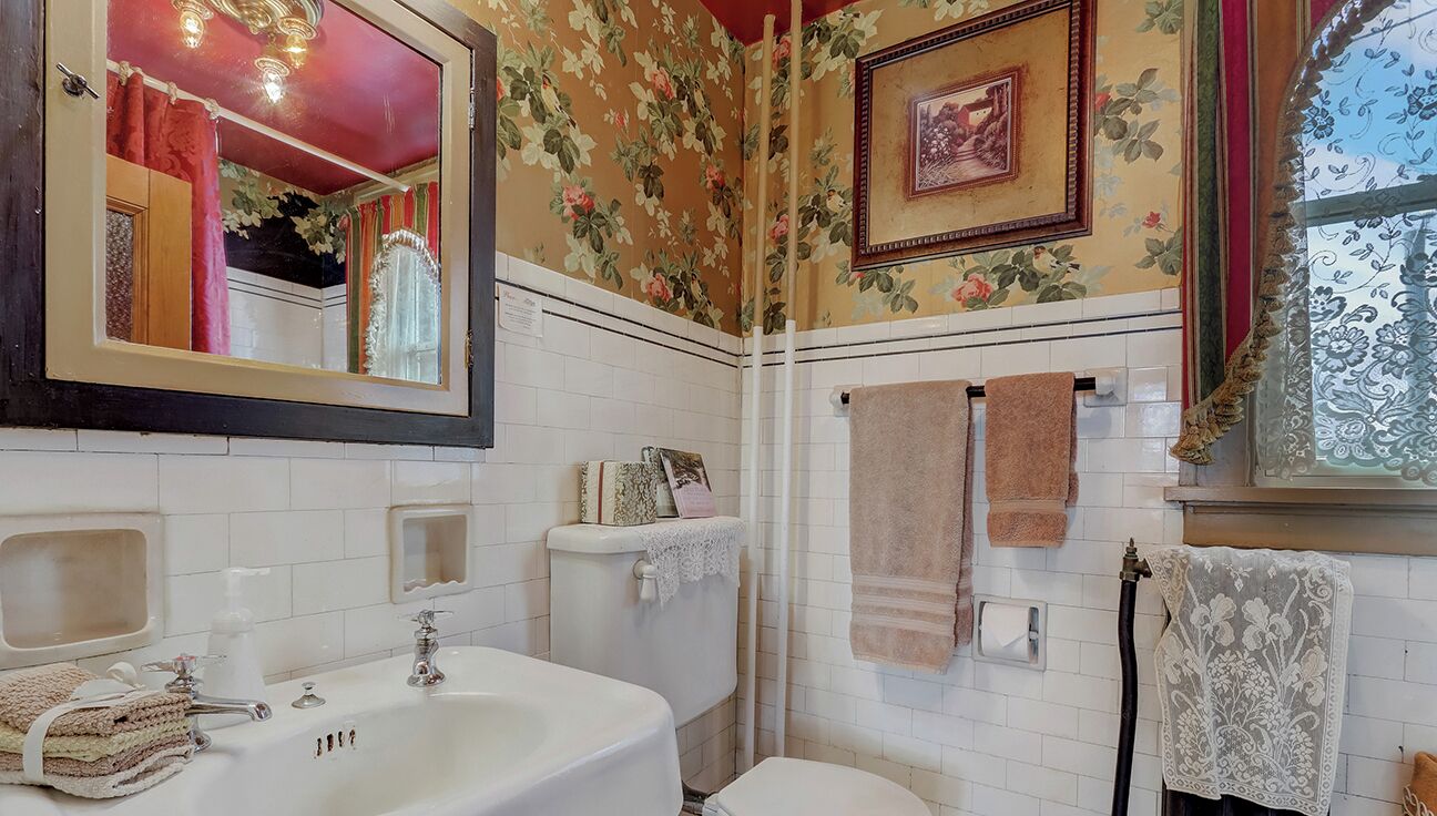 Un baño con lavamanos, espejo y retrete blancos. Presenta papel tapiz con motivos florales y un cuadro enmarcado en la pared.