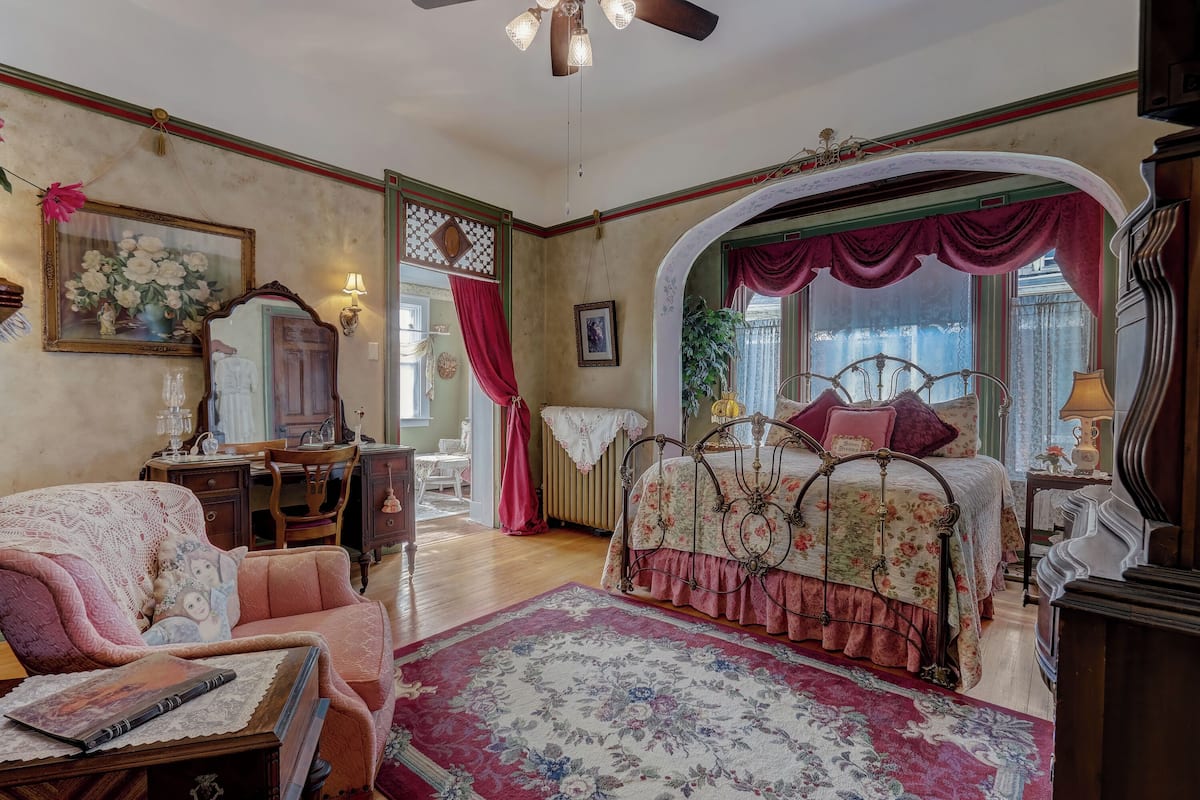 Un dormitorio vintage con una colcha estampada, una silla antigua, un aparador de madera y un espejo grande.