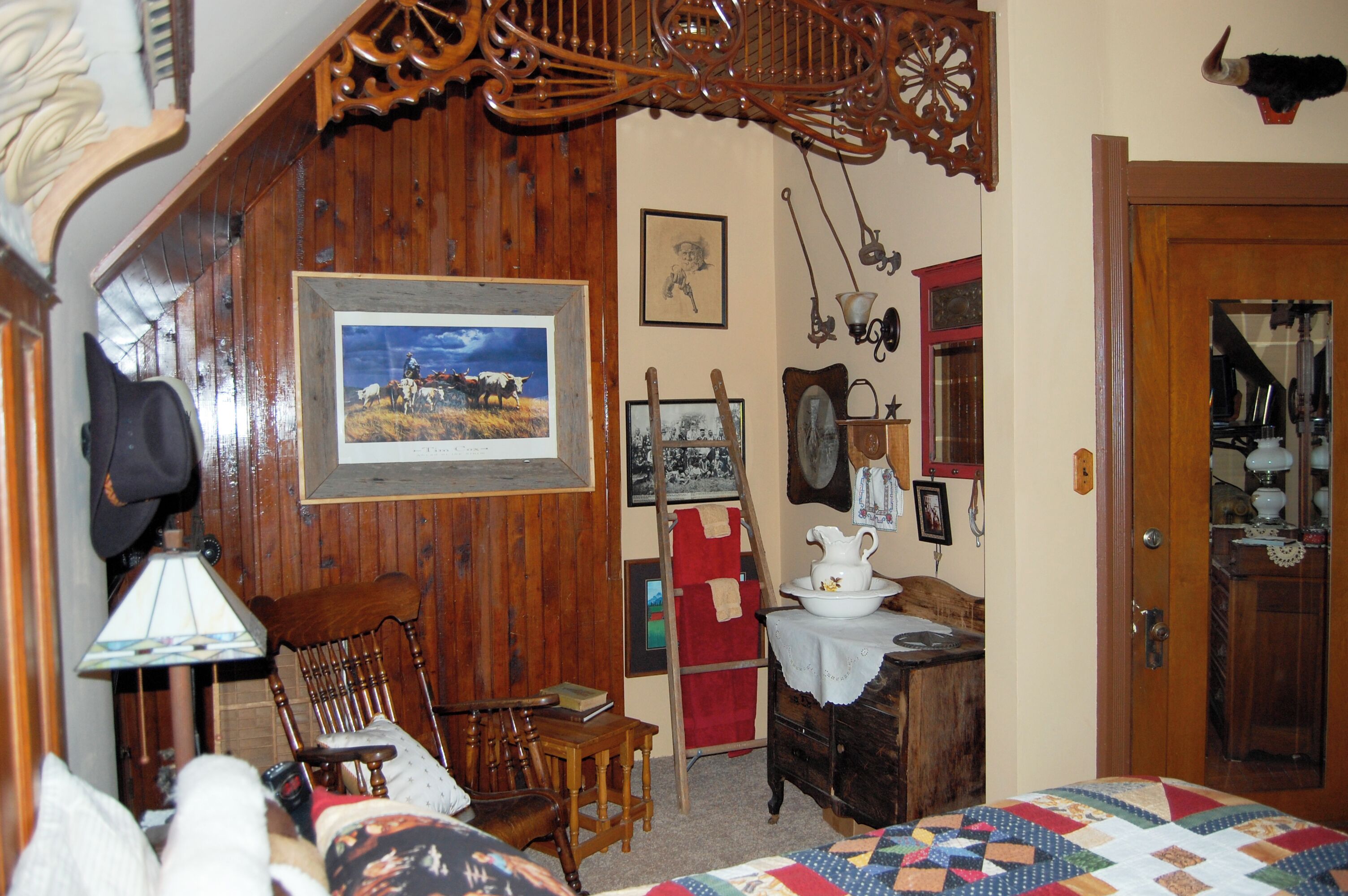 Une pièce rustique aux murs en bois, avec un lit recouvert d’une courtepointe, une chaise en bois, une table sur laquelle se trouve une carafe, et divers éléments décoratifs sur les murs, notamment un tableau encadré et une tête d’animal empaillée.
