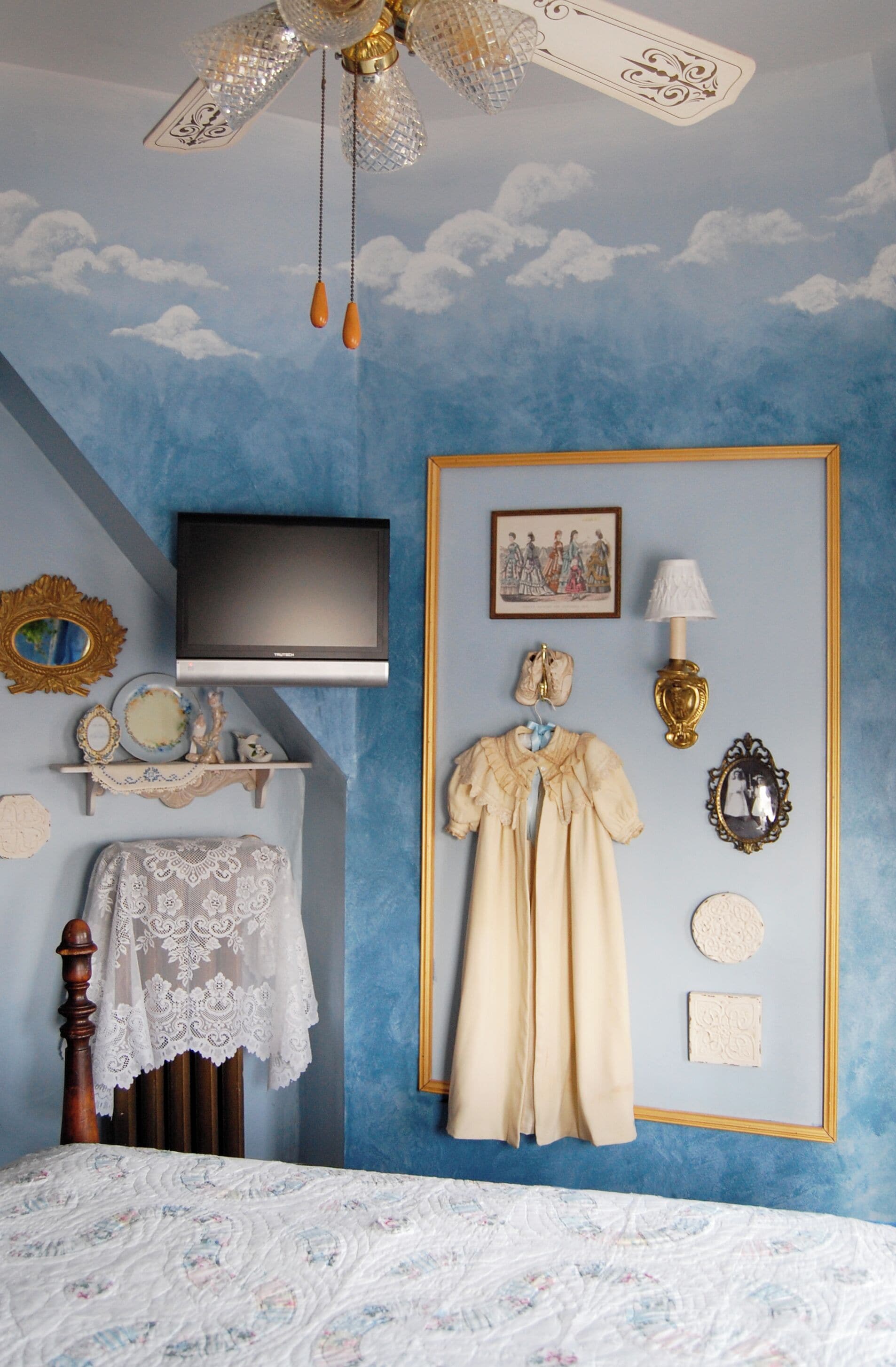 A bedroom with a TV, a dresser, a bed with a lace cover, and a wall with a blue sky mural.