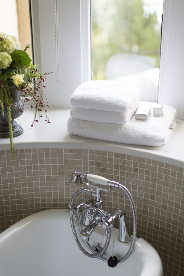 A bathroom with a white bathtub, a window with a view of greenery, and a shelf with white towels and toiletries. - Knockderry Country House Hotel (Helensburgh)