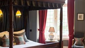 A four-poster bed with a canopy, a grey sofa with cushions, and a chair in a room with striped wallpaper and a window with red curtains.
