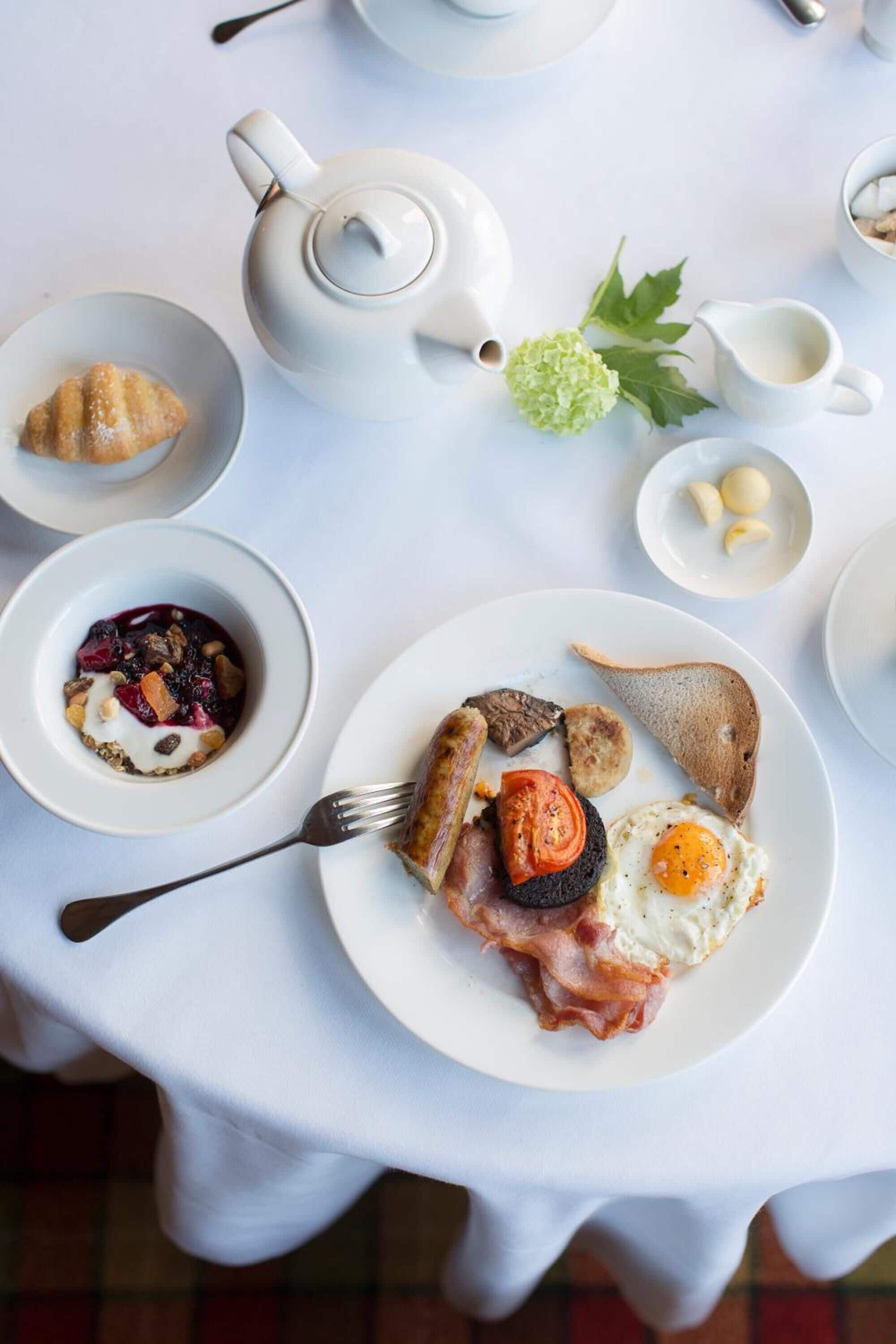 A breakfast setting with a plate of eggs, bacon, sausage, and toast, a bowl of fruit, and a teapot on a white tablecloth.