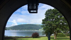 A view through an arched window of a grassy area with a table and chairs, overlooking a lake and a hill.