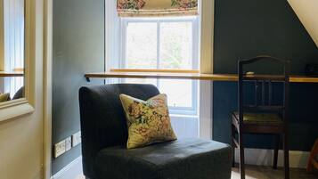 Una habitación acogedora con una pared azul oscuro, una ventana con cortinas florales, una silla de madera y un pequeño otoman con un cojín estampado.