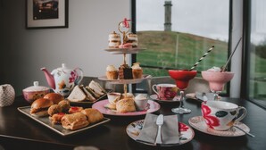 Un juego de té con una bandeja de postres en varios niveles, sándwiches, scones y vistas de una colina a través de una ventana.