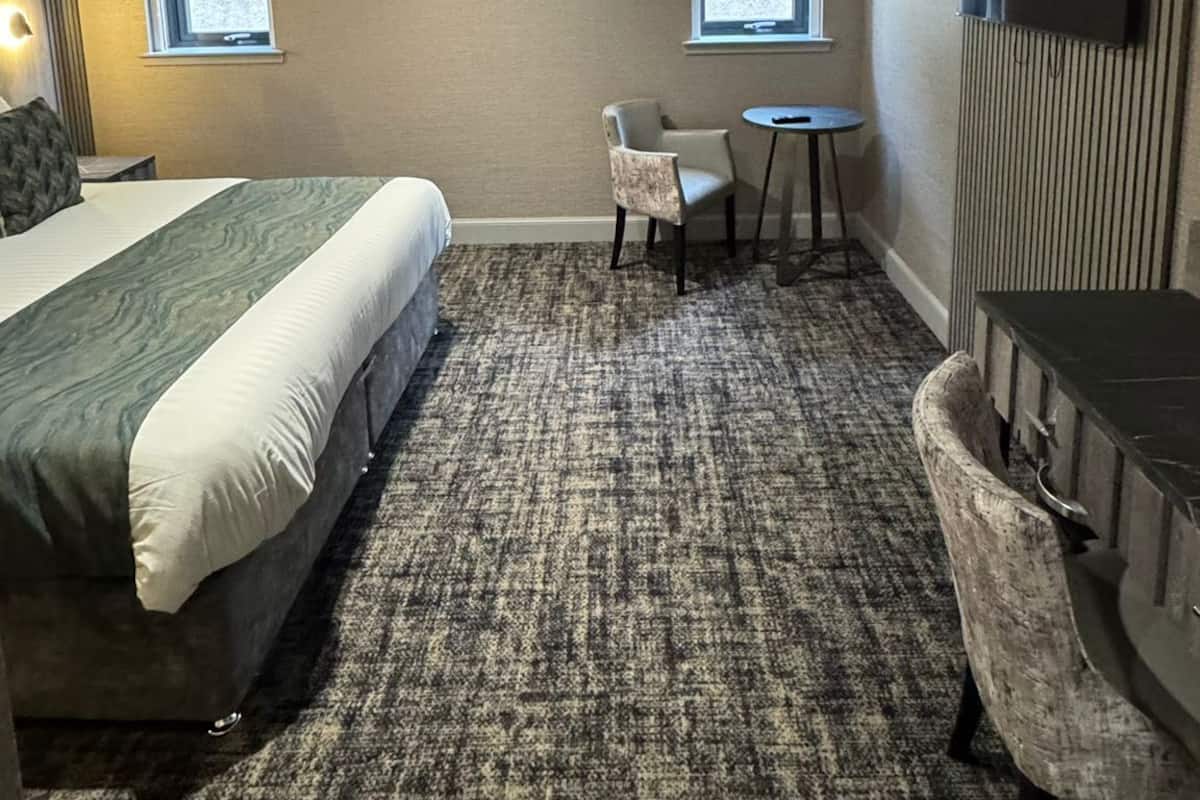 A hotel room with a large bed, a small table, two windows, a TV, and a bathroom vanity.