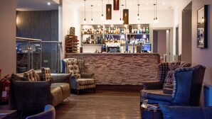 Zona de bar con pared de ladrillo, diversas opciones de asientos que incluyen sofás y sillones, y una variedad de botellas y vasos en estanterías.