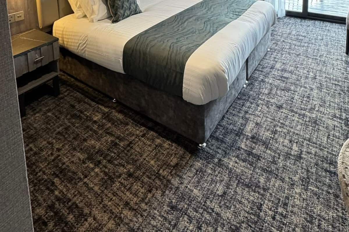 A modern hotel room with a large bed, a headboard with vertical lines, a nightstand, a wall-mounted light, and a view through a large window.