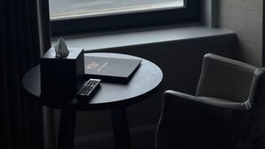 Una habitación con vistas al mar a través de un ventanal grande, una pequeña mesa redonda con una caja de pañuelos y un control remoto, y una silla.
