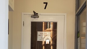 Une porte blanche avec le numéro 7 au-dessus, une fenêtre donnant sur un immeuble et un interphone à l’intérieur.