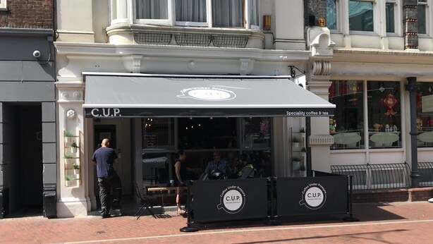 Une devanture de magasin avec un auvent noir et une enseigne indiquant : « C.U.P. – Café et thé de spécialité ».
