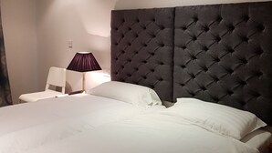 Un lit bien fait, avec du linge de lit blanc, une tête de lit capitonnée de couleur sombre, une chaise blanche et une lampe de table avec un abat-jour foncé.