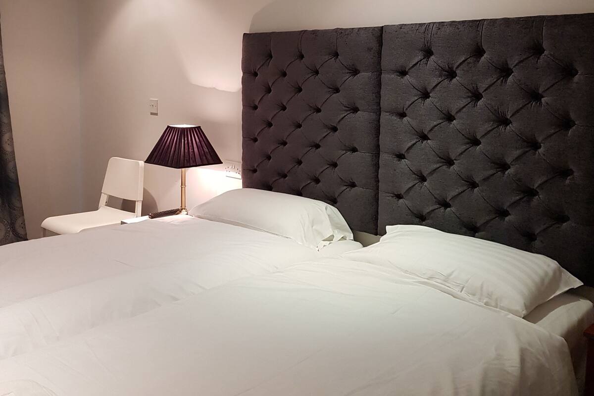 A neatly made bed with white linens, a dark tufted headboard, and a bedside lamp.