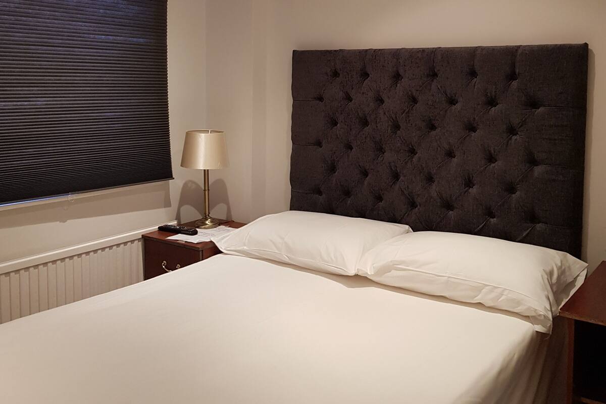 A neatly made bed with white linens, a dark tufted headboard, a bedside lamp, and a window with blinds.