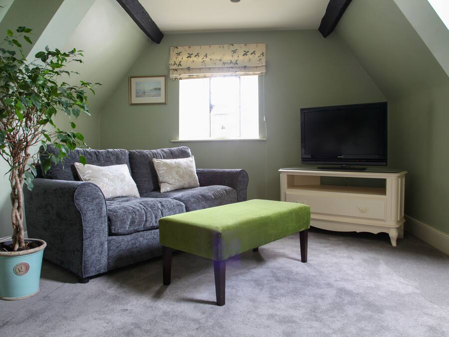 A cozy room with a grey sofa, a green ottoman, a potted plant, a television on a stand, and a window with a floral valance.
