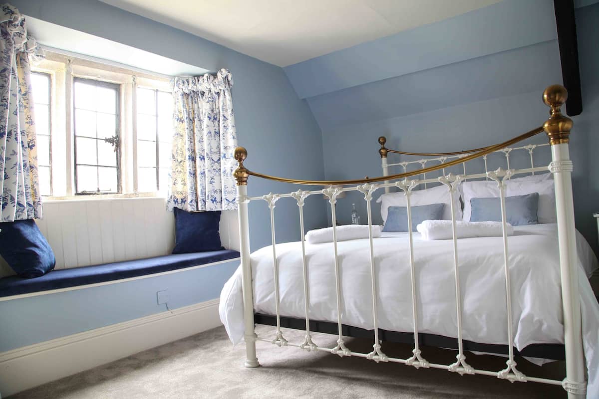 Quarto com cama de dossel branca, janela com cortinas azuis e um banco embutido com almofadas.