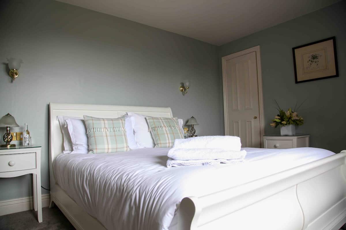Cama bem arrumada com roupa de cama branca e várias almofadas, mesa de cabeceira com abajur e criado-mudo com um vaso de flores.