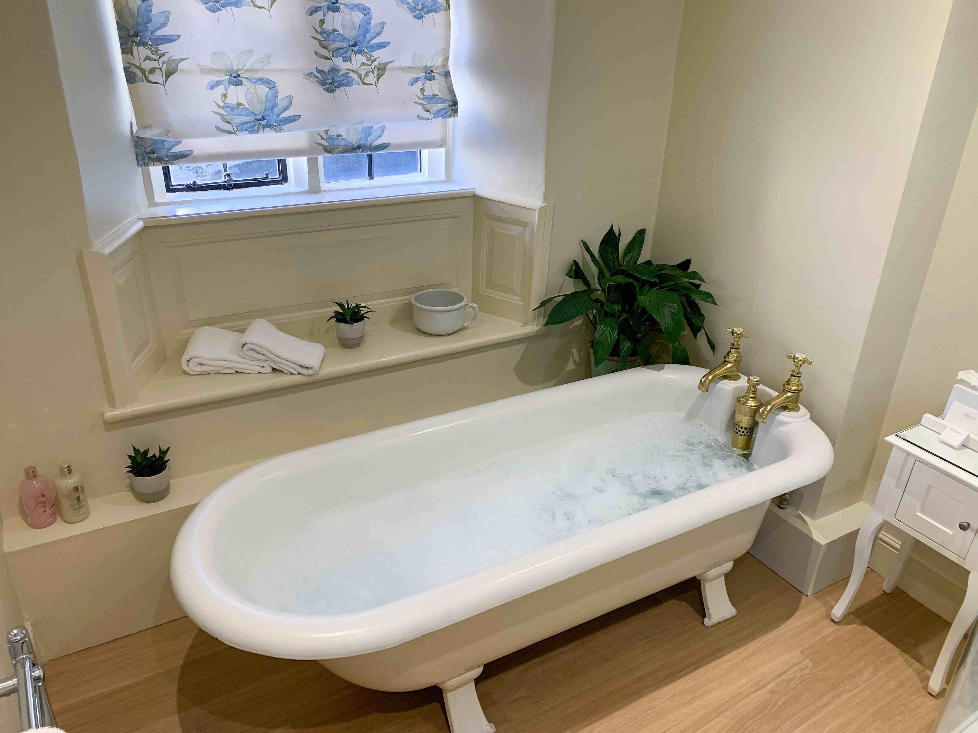 Un baño con una bañera independiente, una ventana con cortinas florales, un estante con artículos de aseo y una planta en maceta.