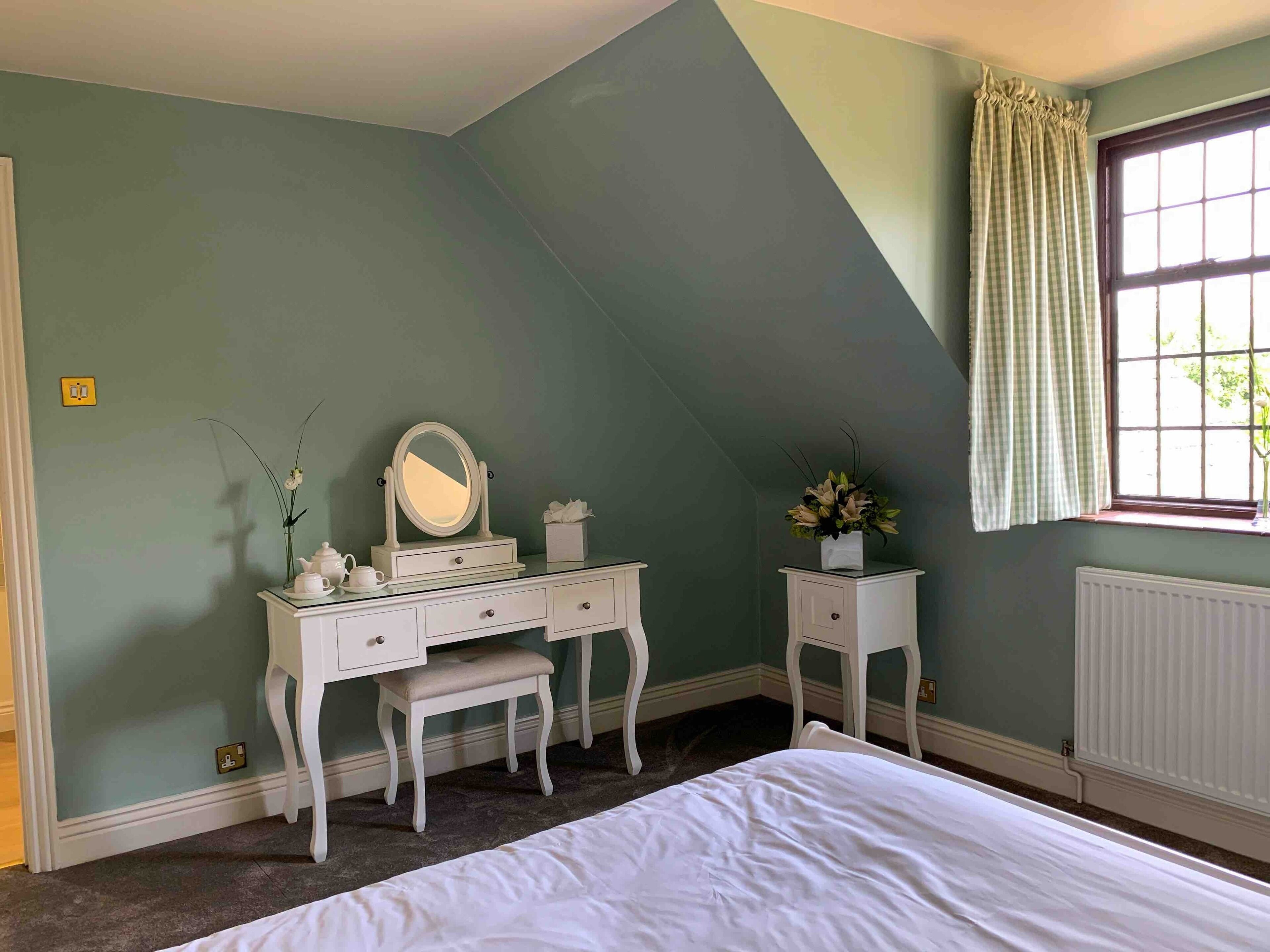 A bedroom with a bed, a dressing table with a mirror, a small stool, and a window with curtains.