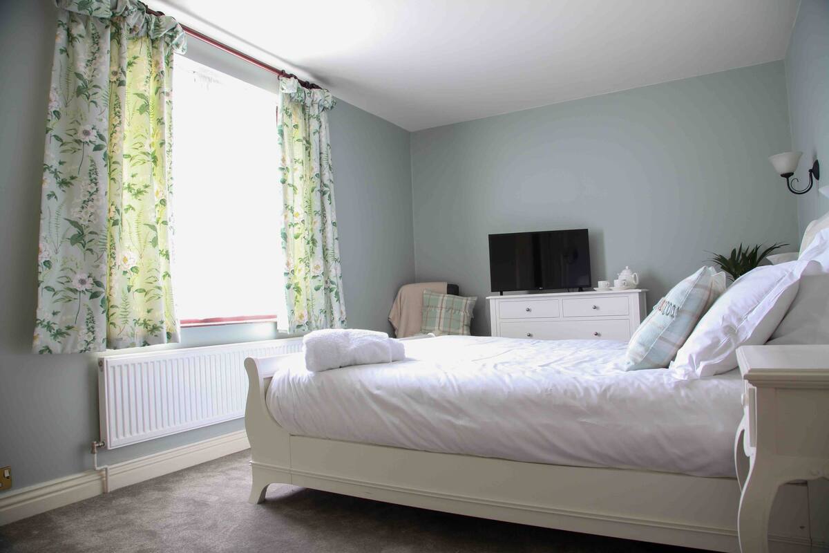Quarto com cama, penteadeira, televisão e janela com cortinas.