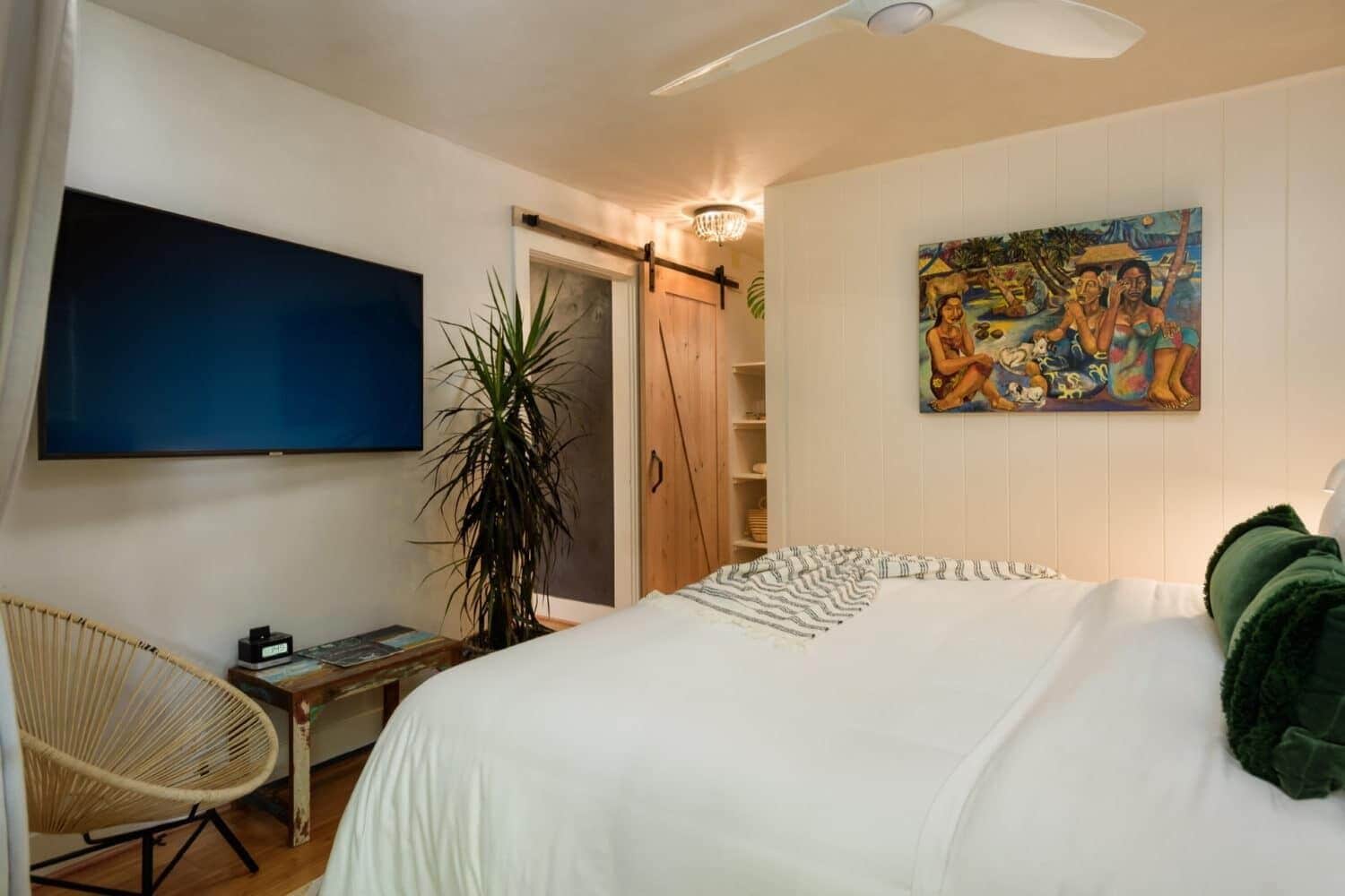 A bedroom with a bed, a desk, a chair, a TV, a plant, and a painting on the wall.