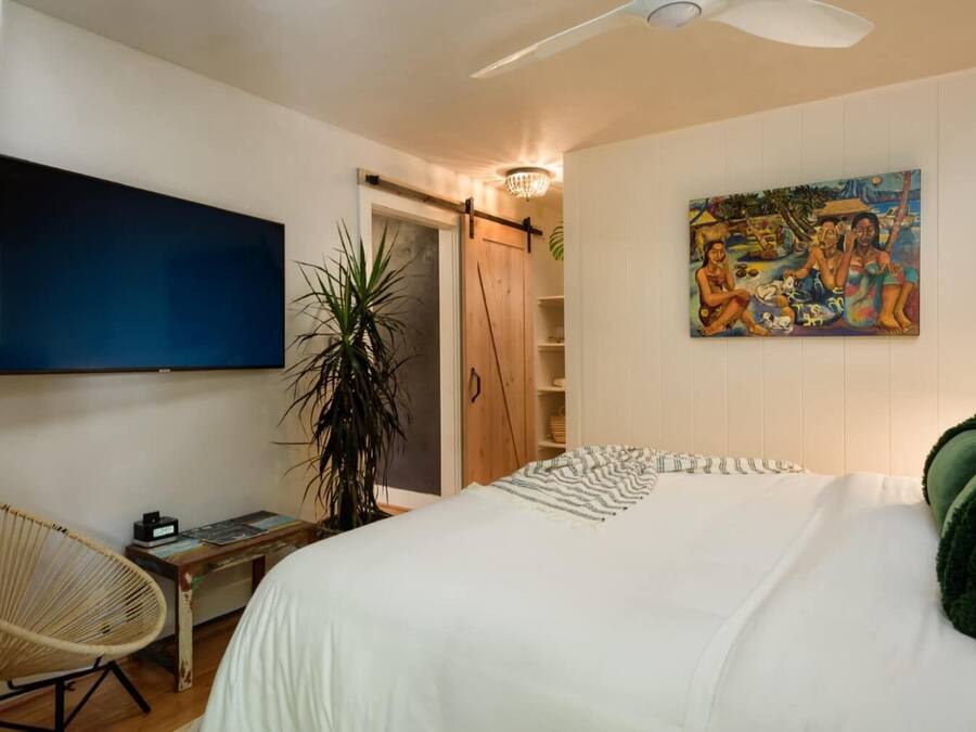Un dormitorio con cama, escritorio, silla, televisión, planta y un cuadro en la pared.