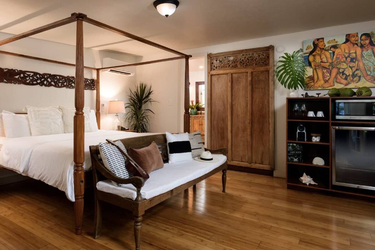 Ein Bett mit vier Säulen und Holzbaldachin, eine Holzbank mit Kissen, eine Wand-Klimaanlage, ein Topfpflanze, ein Holzschrank und ein Gemälde an der Wand.