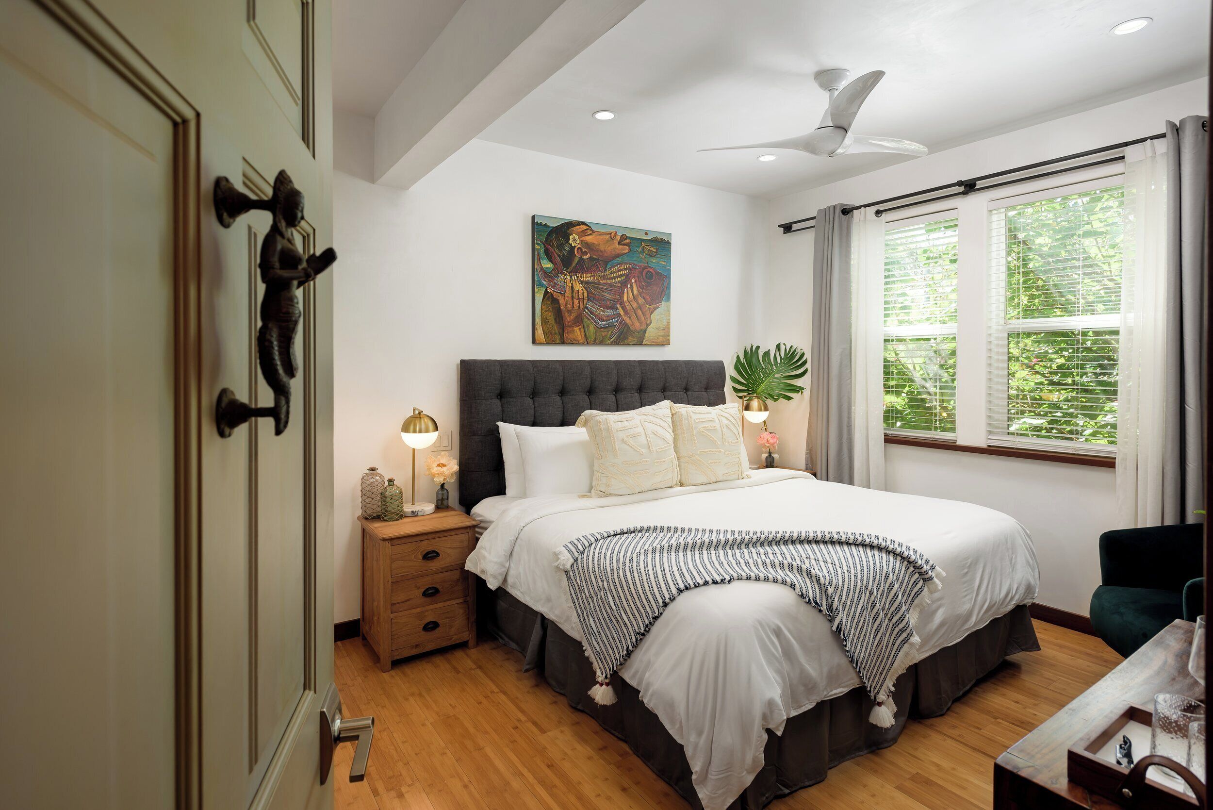 A bedroom with a large bed, a nightstand, a ceiling fan, a window with curtains, and a painting on the wall.