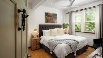 A bedroom with a large bed, a nightstand, a ceiling fan, a window with curtains, and a painting on the wall.