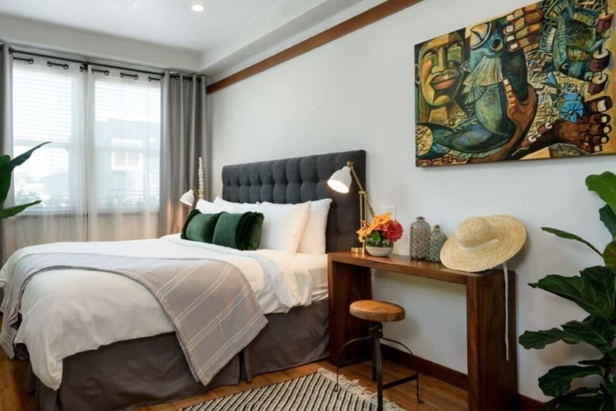 Ein Schlafzimmer mit Bett, Nachttisch, Schreibtisch, Stuhl, einem Gemälde und einer Topfpflanze.