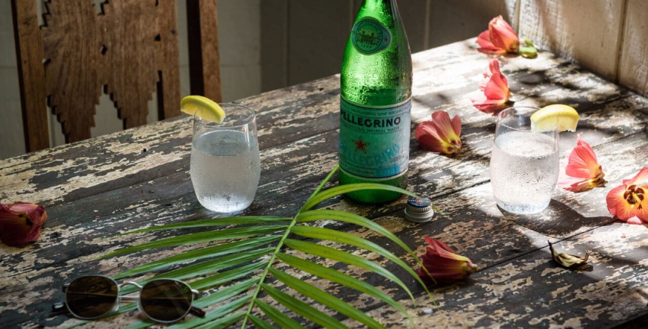 A rustic wooden table with two glasses of water, a green bottle labeled 'ELLECRINO', sunglasses, and scattered flowers.