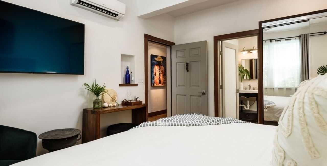 A hotel room with a bed, a TV, a desk with a lamp, a door, and a bathroom visible through an open door.