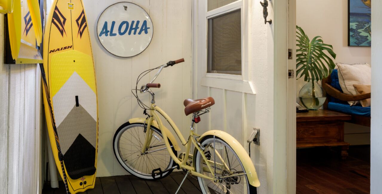 Una bicicleta antigua con asiento de cuero marrón, una tabla de surf y un letrero en la pared que dice 'ALOHA'.
