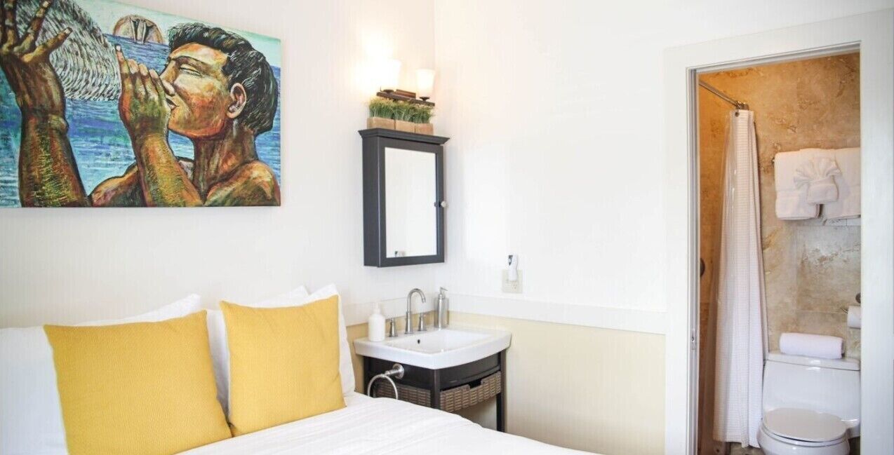 A hotel room with a bed, a small bedside table with a lamp, a painting on the wall, a bathroom with a shower, and a toilet.
