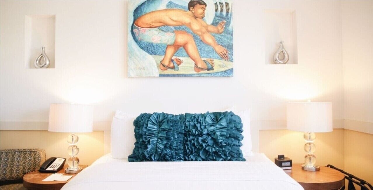 Habitación de hotel con cama, dos lámparas de noche, un cuadro de un surfista y una almohada decorativa.