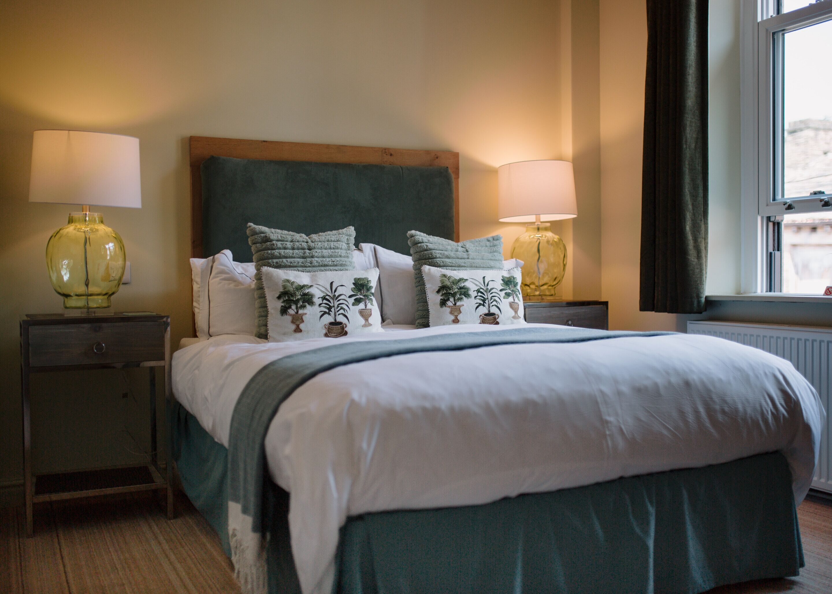A neatly made bed with a green bedspread and multiple pillows, including two with a palm tree design. There are two bedside lamps on wooden nightstands.