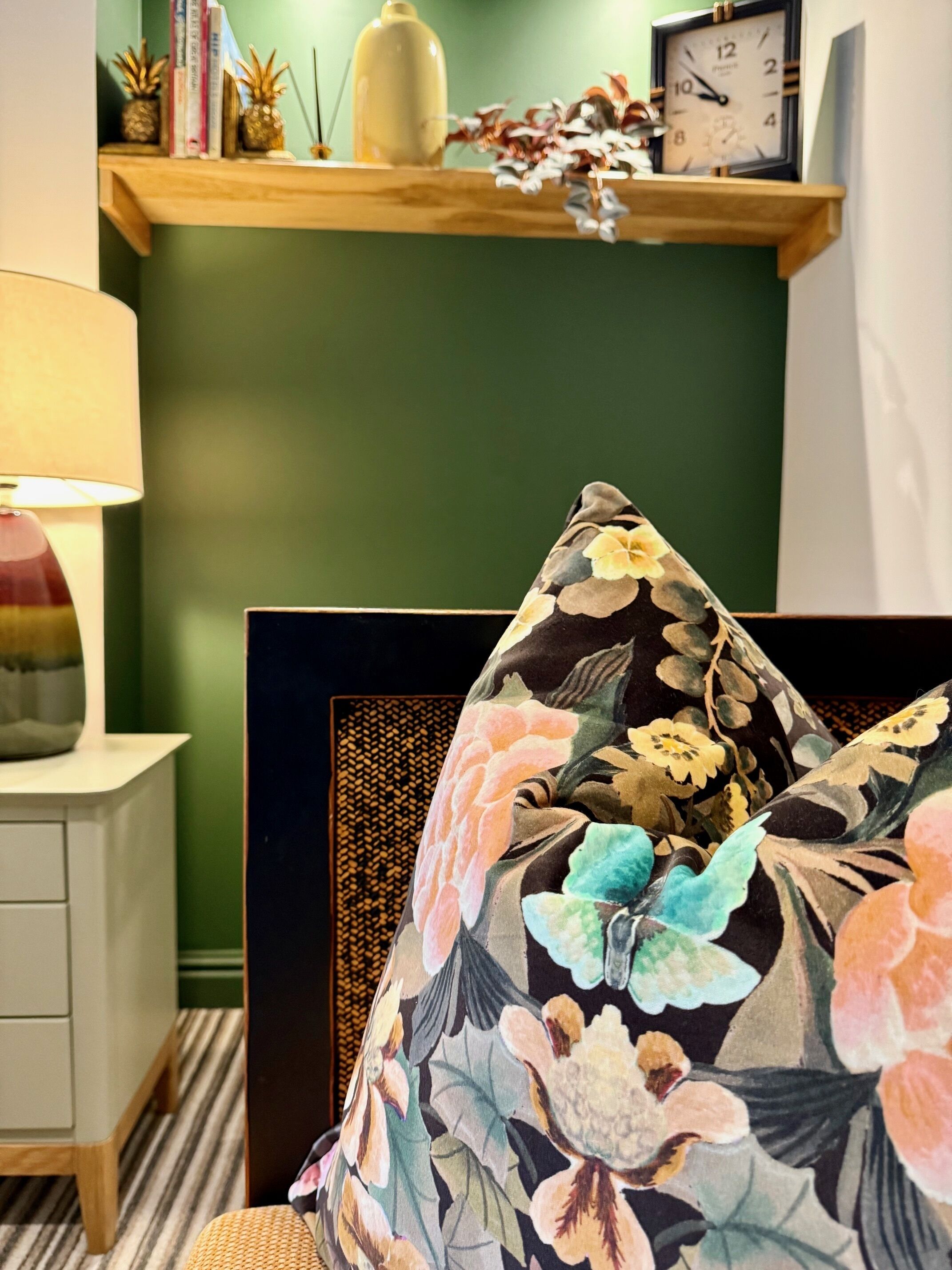 Ein Zimmer mit grüner Wand, einem hölzernen Regal mit Büchern und Dekoration, einer Uhr und einem blumigen Kissen auf einem Stuhl.