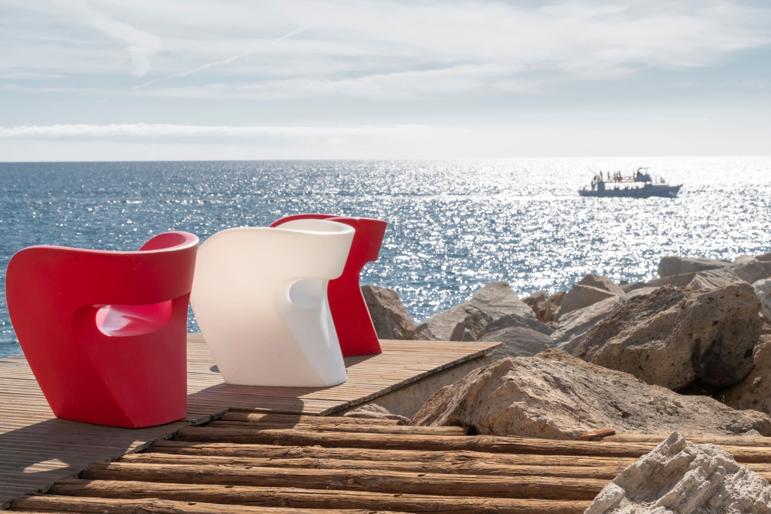 A wooden deck with two chairs, one red and one white, overlooking a rocky shoreline and a boat on the water.
