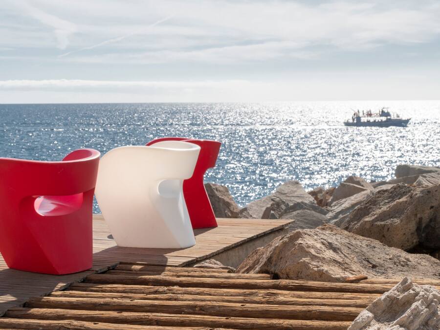 Terraza de madera con dos sillas, una roja y otra blanca, con vista a una costa rocosa y un bote en el agua.
