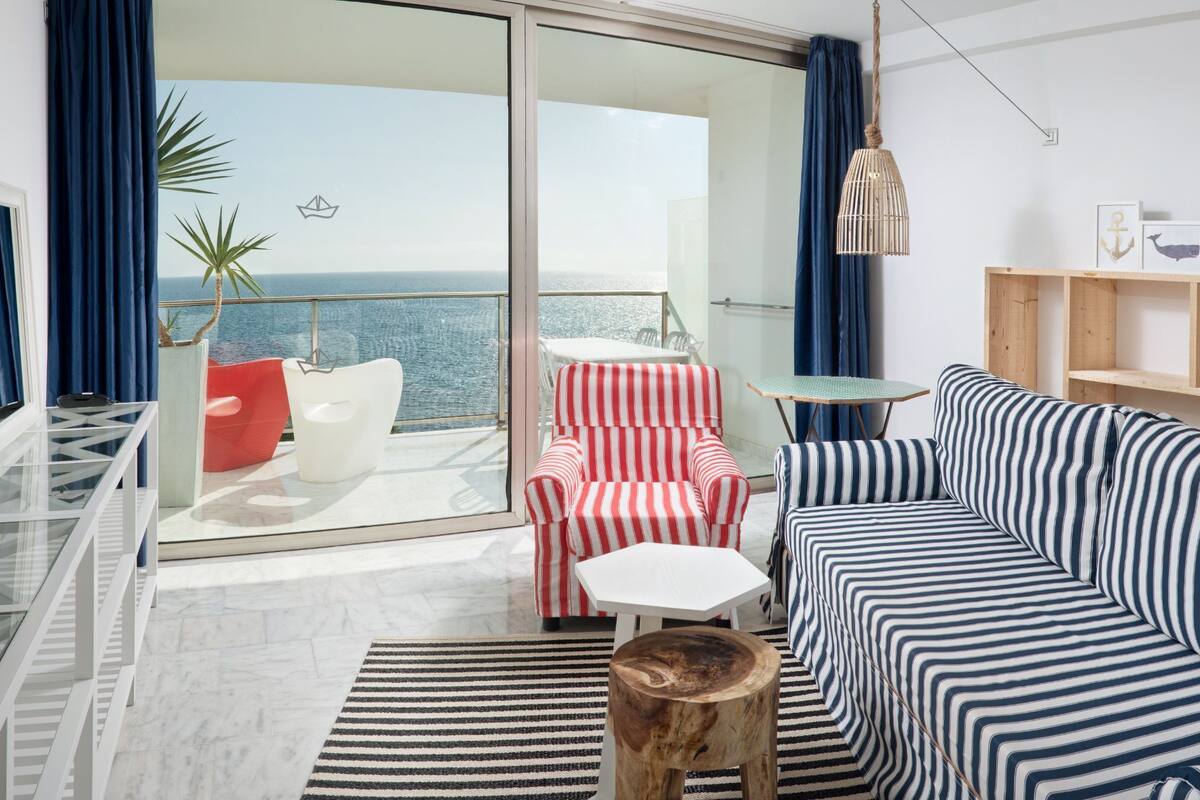 Sala de estar moderna com um sofá, uma cadeira, uma mesinha e vista para o mar através de uma porta de vidro.