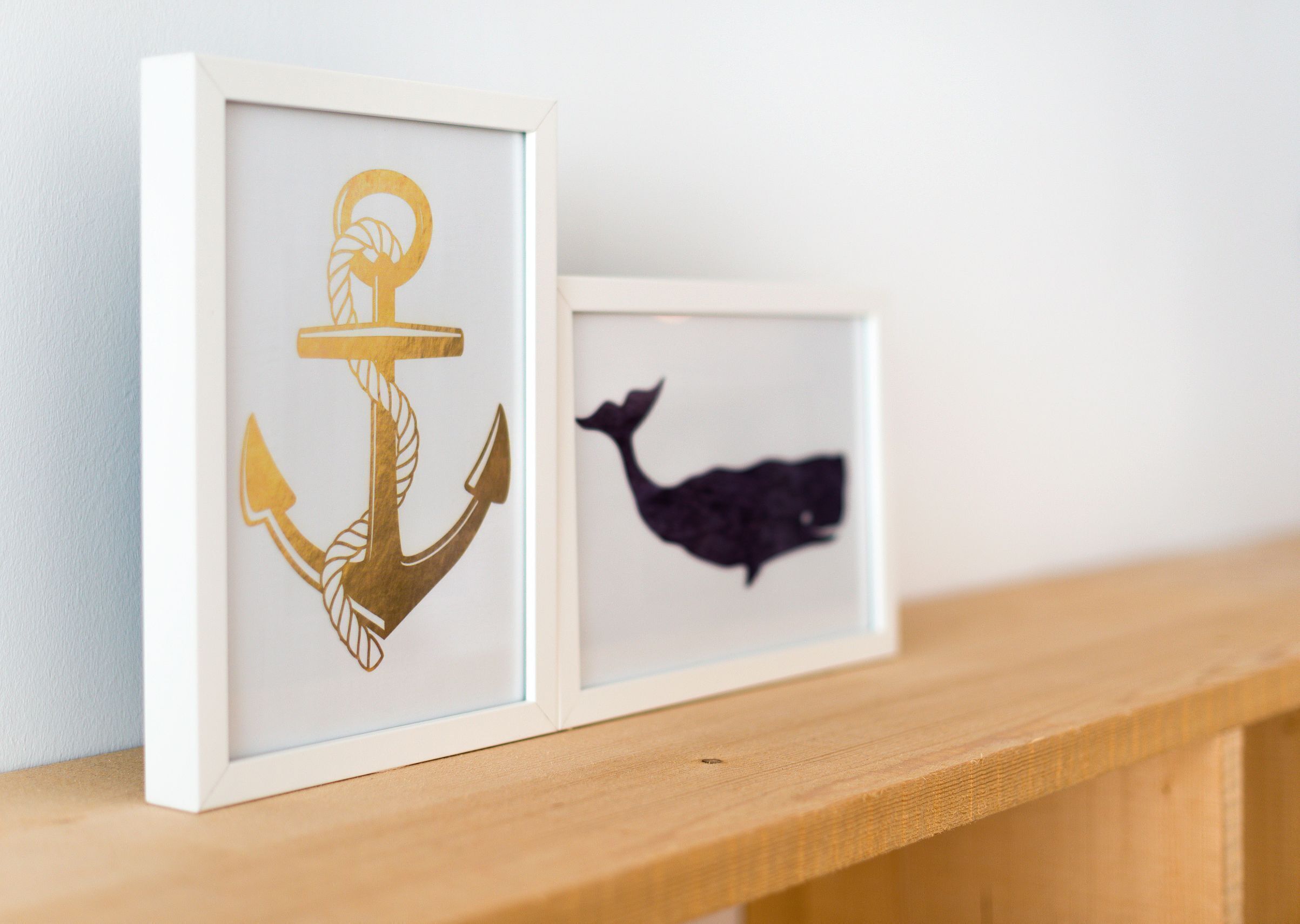 Two framed artworks on a shelf: one with a golden anchor and the other with a silhouette of a whale.