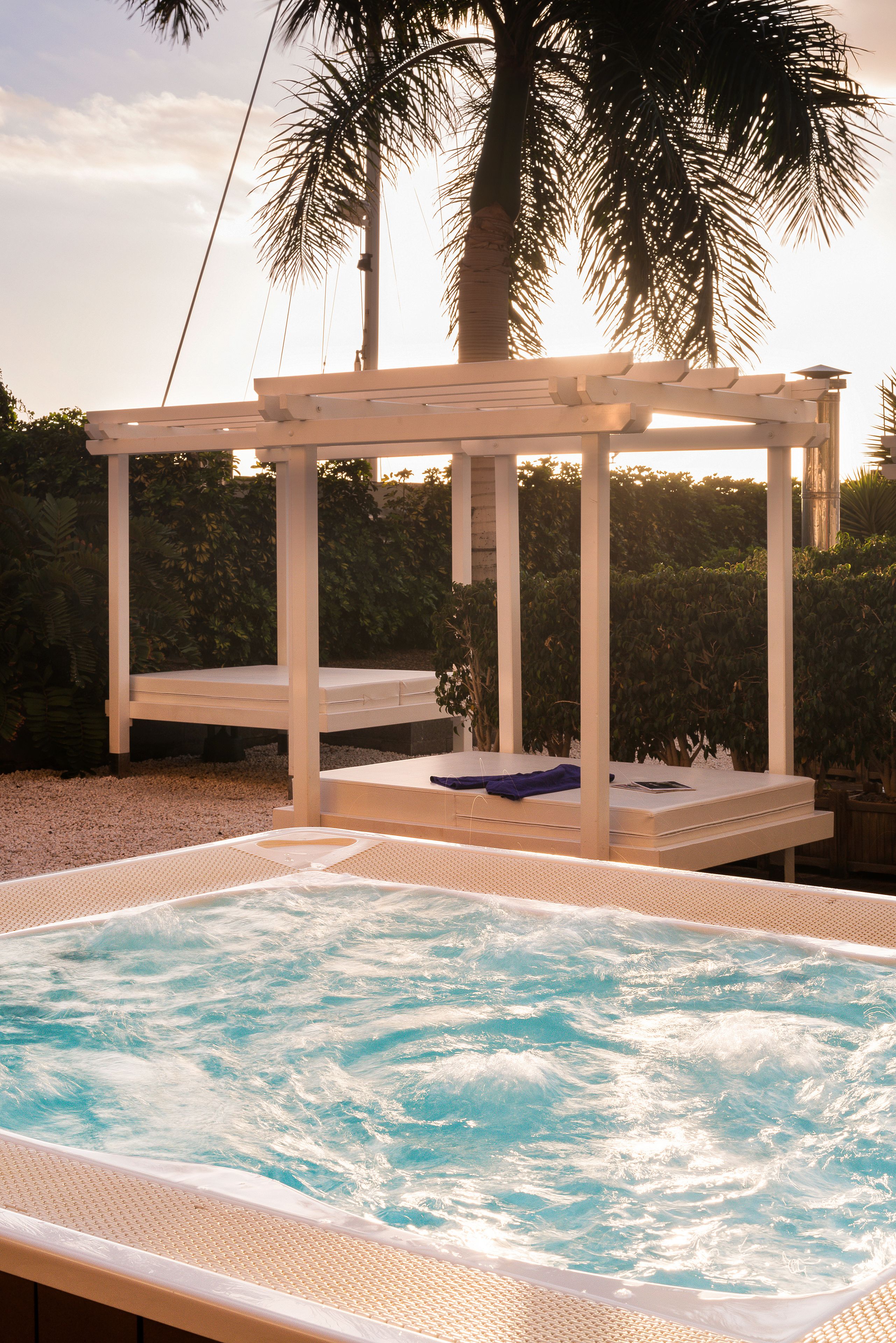 A hot tub with a wooden pergola structure, surrounded by palm trees and a hedge.