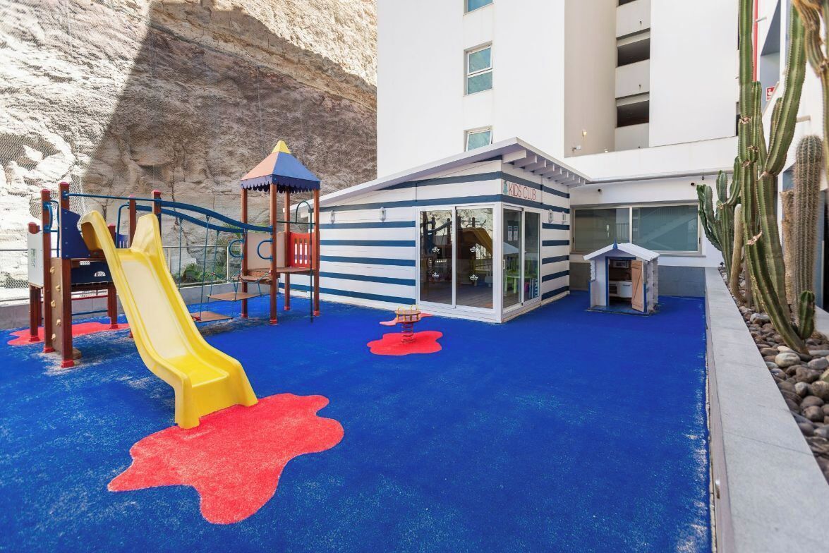 A children's play area with a slide and climbing structures, adjacent to a building with a 'Kids Club' sign.