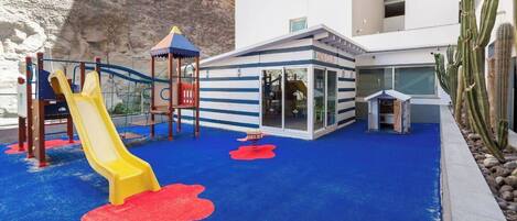 A children's play area with a slide and climbing structures, adjacent to a building with a 'Kids Club' sign.