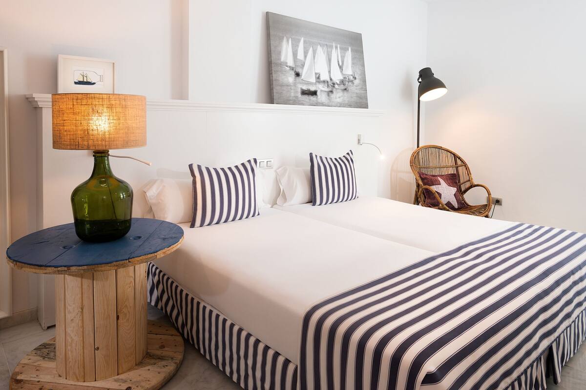 Uma cama bem arrumada com roupa de cama listrada, uma mesa redonda de madeira com um vaso verde, uma cadeira de vime e um abajur fixado na parede.