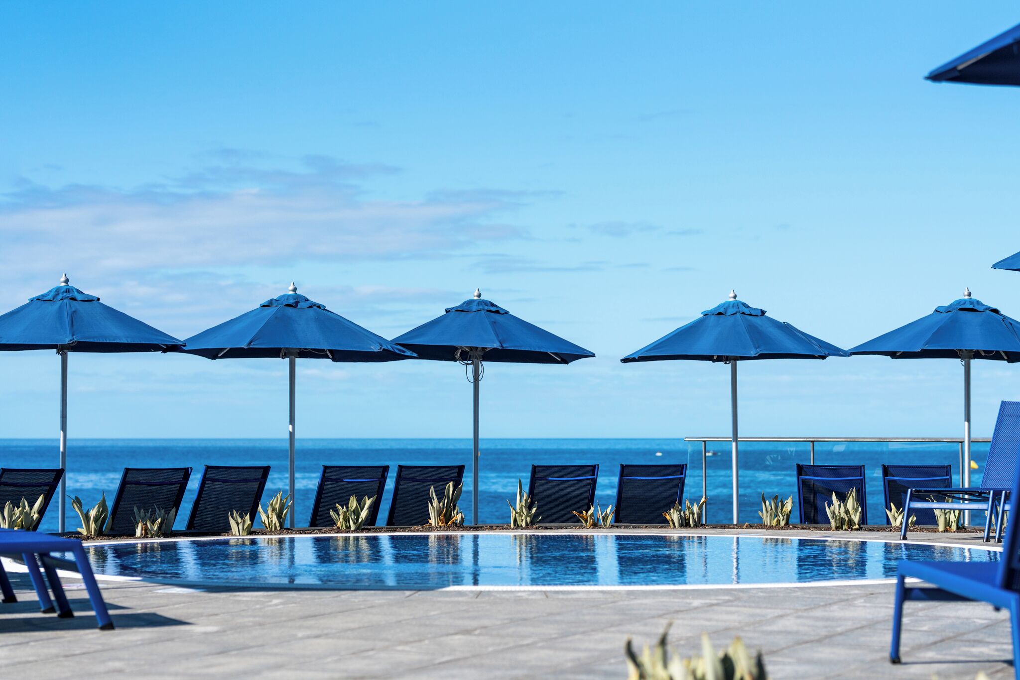 A poolside area with blue umbrellas and lounge chairs facing the ocean.