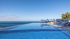 Una piscina infinita con vista al océano, rodeada de sillones de descanso azules y sombrillas.