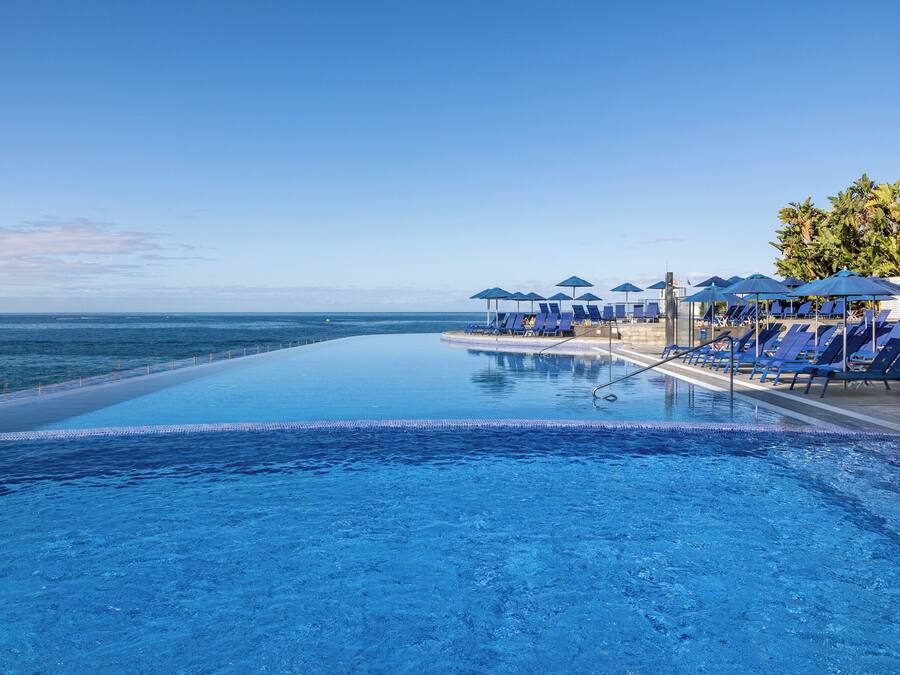 Una piscina infinita con vista al océano, rodeada de sillones de descanso azules y sombrillas.